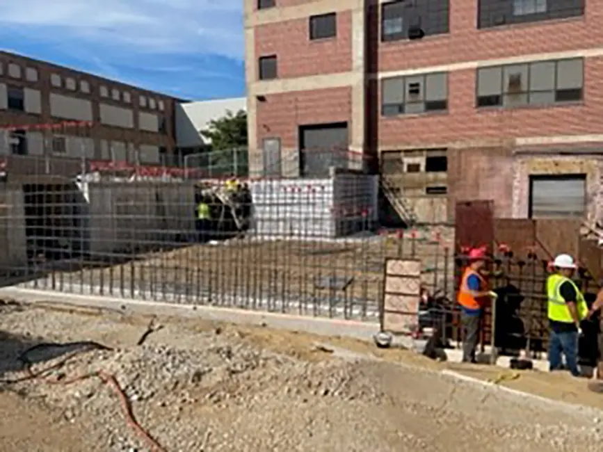 Construction site for addition onto existing brick building. Site includes concrete foundation with rebar structures. Workers with safety vests are visible. Surrounding area includes gravel and construction materials with additional buildings and clear blue sky in the background.