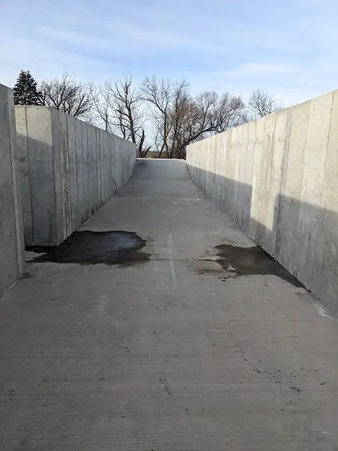 Concrete ramp with high retaining walls on both sides, leading upward toward an open area with trees visible in the background. The surface of the ramp has some wet patches and marks.